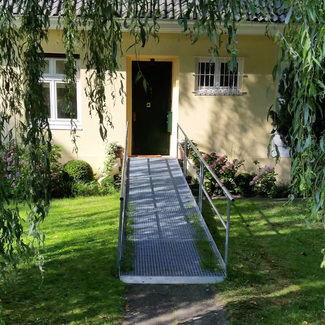Rampe für Rollstühle im Garten auf Rasen. Führt zur Haustür eines gelb gestrichenen Einfamilienhauses.