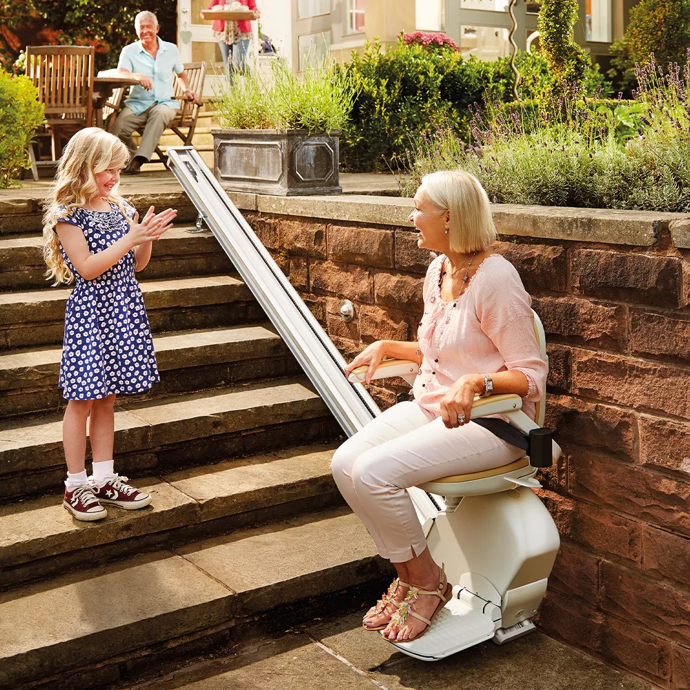 Eine Frau sitzt auf einem Treppenlift, der die Terrasse mit dem Garten verbindet. Die Enkeling schaut zu. Rhein-Mosel Lifte bietet Treppenlifte für den Außenbereich an.