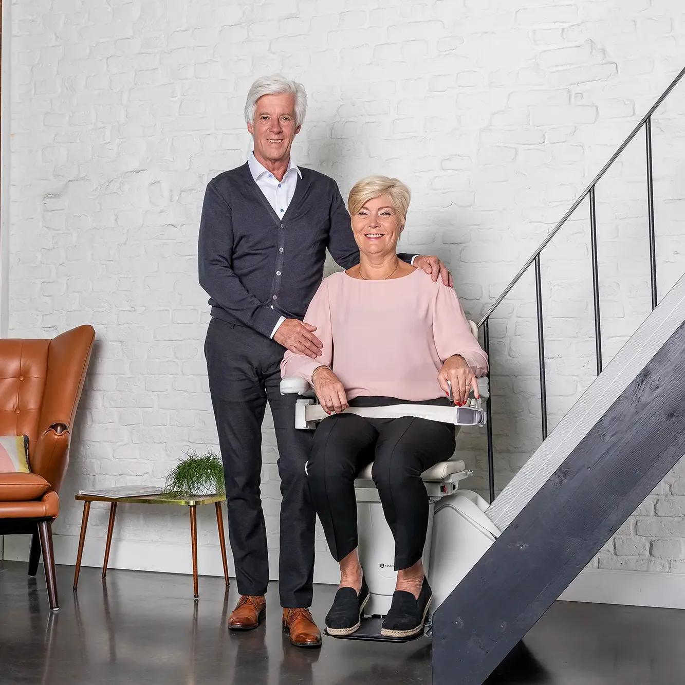 Treppenlift von der Firma Handicare. Eine Frau sitzt im Treppenlift und Ihr Mann steht neben ihr. Rhein-Mosel Lifte führt Maschinen von Handicare.